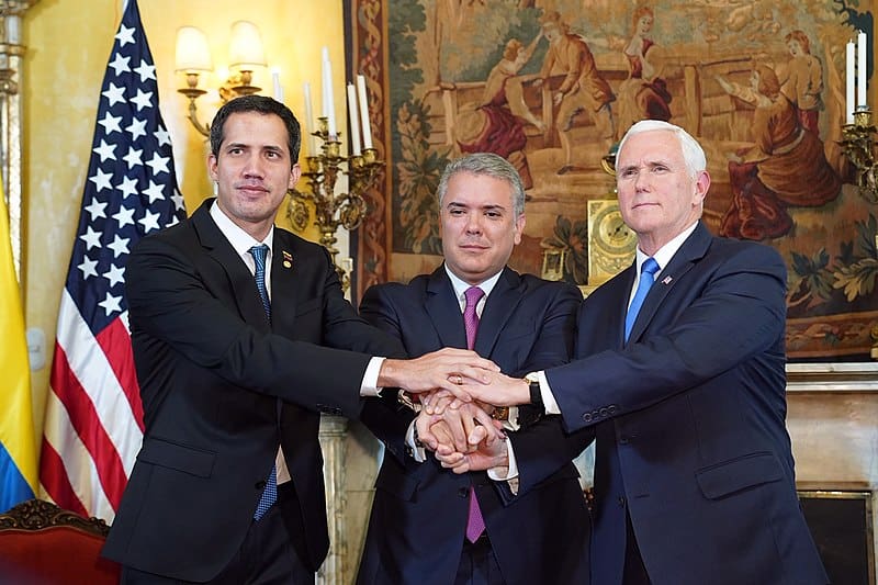 Three men in suits shake hands in front of an american flag.
